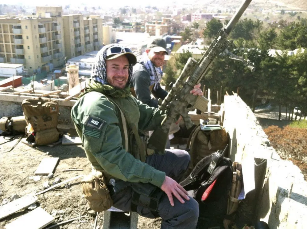 Sonny smiling on a rooftop overlooking a city, rifle at hand