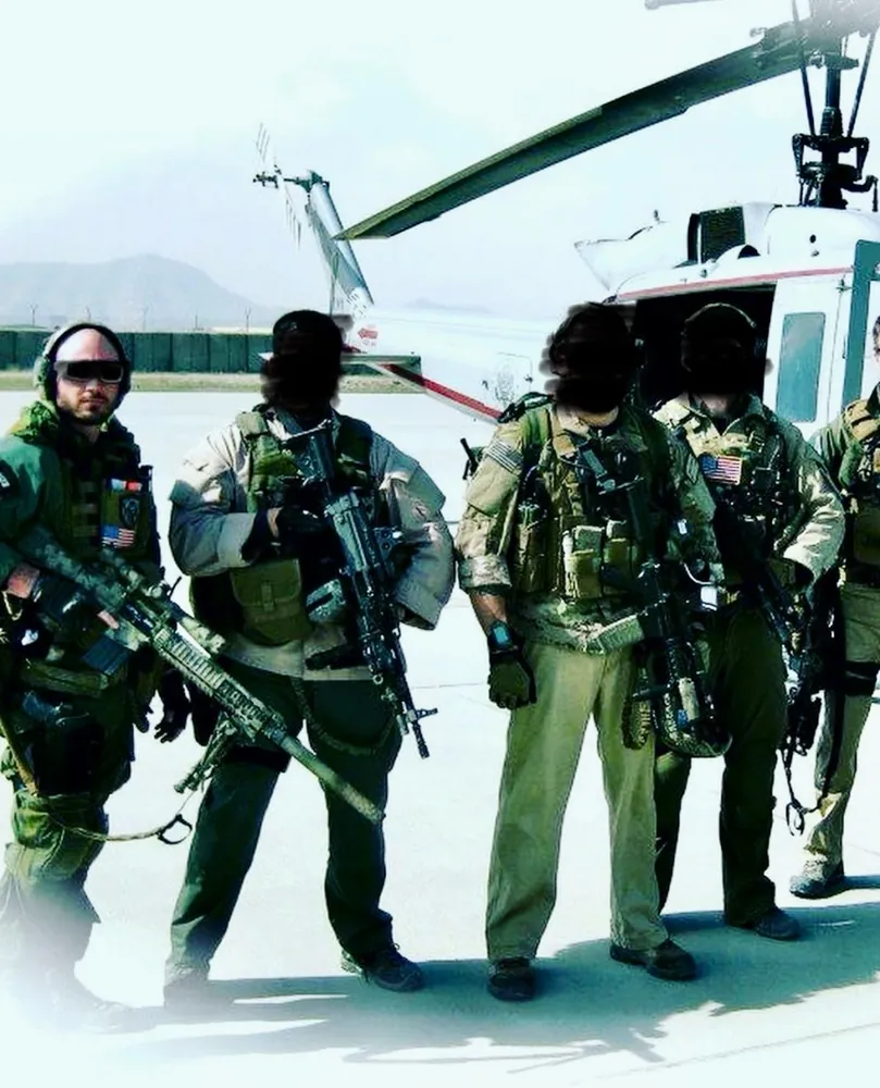A team in gear standing in front of a helicopter on a flight line, other faces blurred